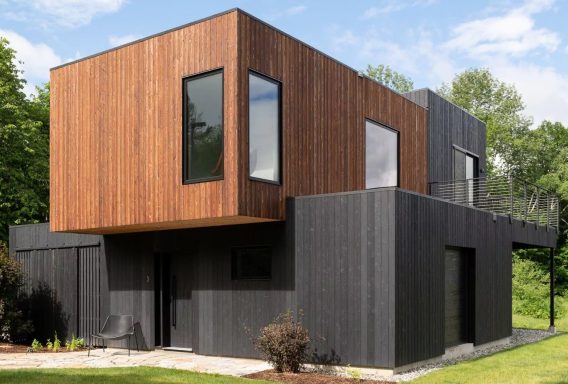 Modernes Haus mit Holzverkleidung und großen Fenstern, umgeben von Grün.